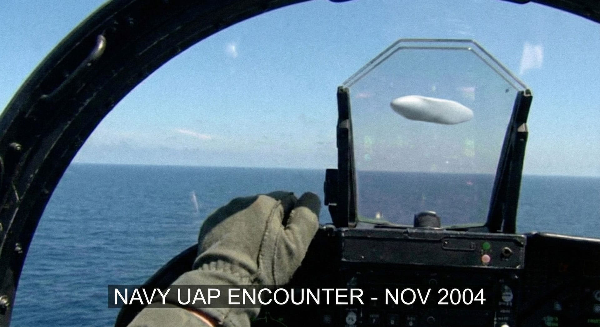 View from inside a US Navy fighter jet cockpit during the 2004 Nimitz encounter, showing a pilot's gloved hand and a white "Tic Tac" shaped UAP hovering over the ocean with a timestamp overlay.